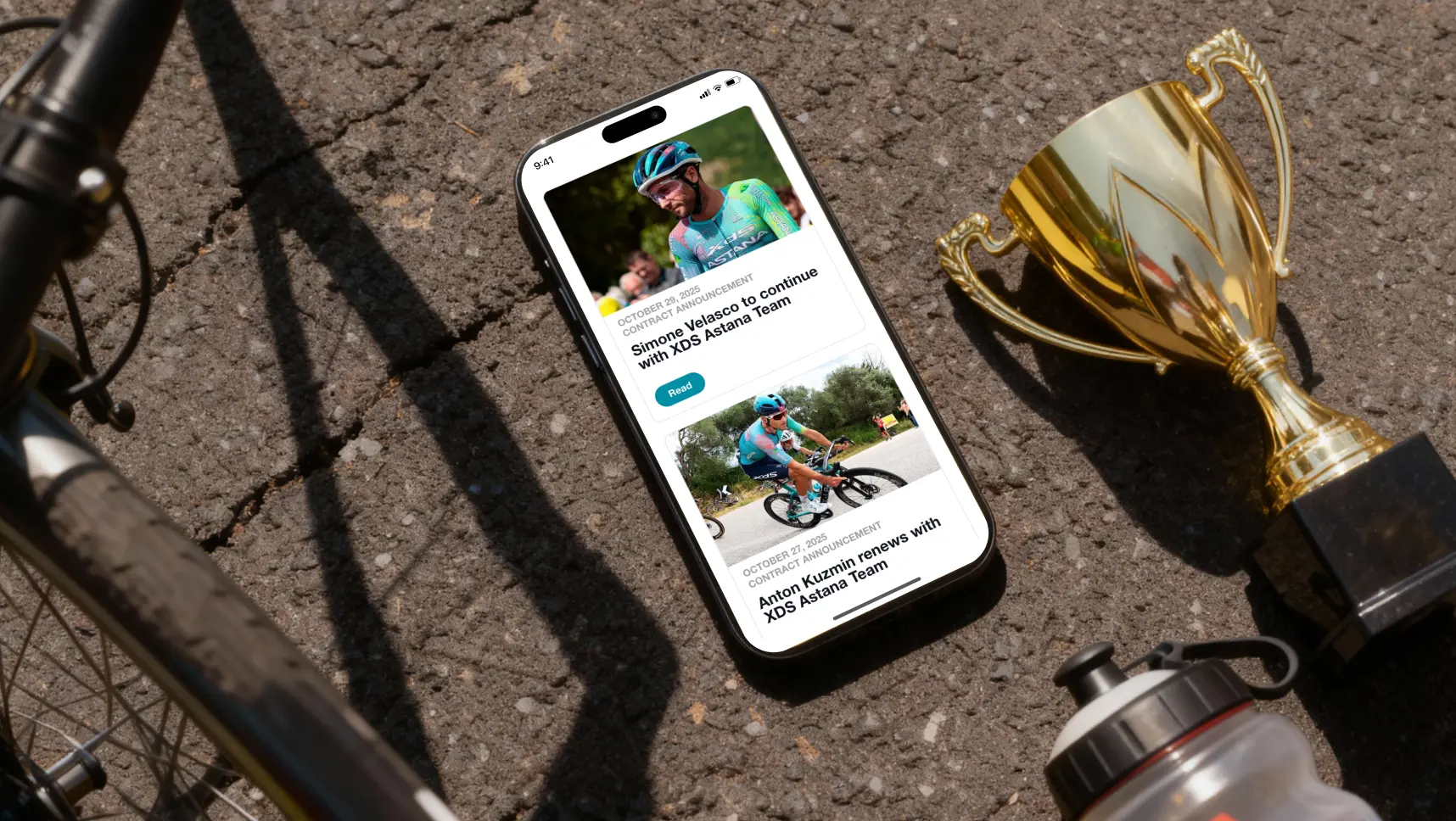 Cycling news feed displayed on a smartphone next to a trophy and bike gear