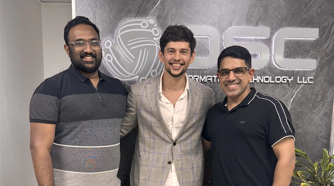 Three men standing side by side and smiling in an office, in front of an illuminated wall sign reading "DSC Information Technology LLC".