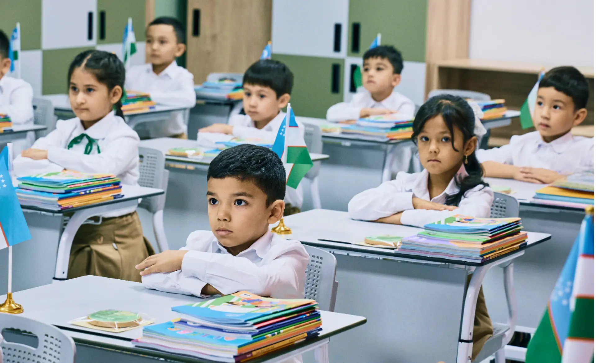 Primary school students in uniforms sitting at desks with Uzbekistan flags and textbooks in a modern classroom.